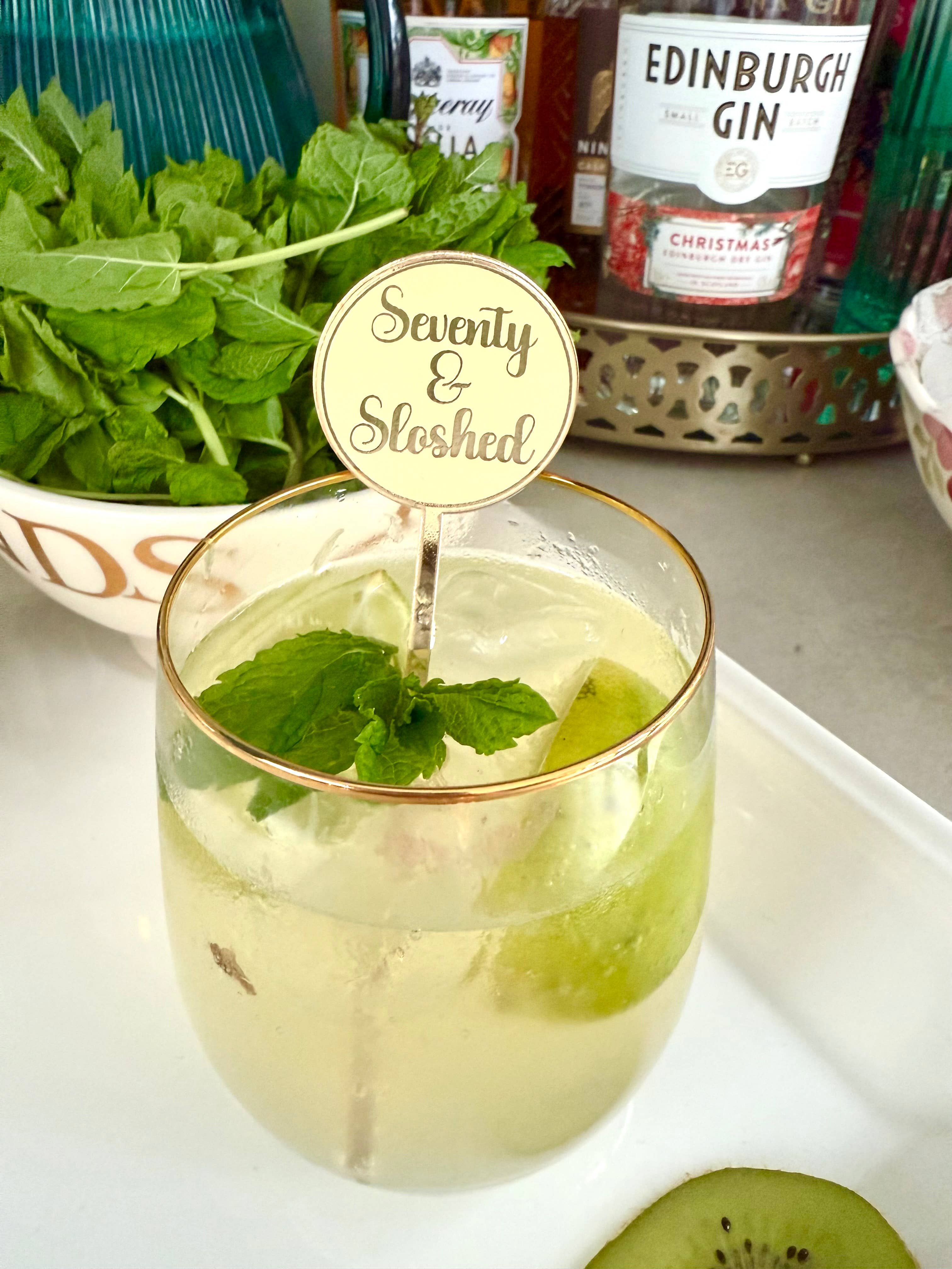 Glass of cocktail with mint leaves and a 'Seventy & Sloshed' stirrer, surrounded by bottles and ingredients on a countertop.